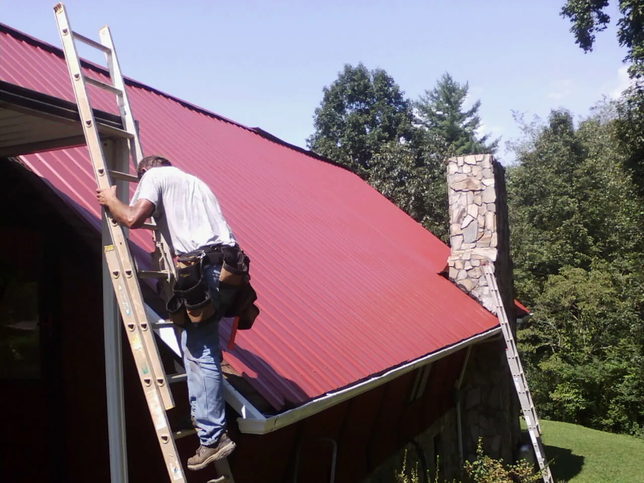 Roofer on a ladder beside a red metal roof and stone chimney — Soffit & Fascia Repair by Roofing Brothers in Russellville